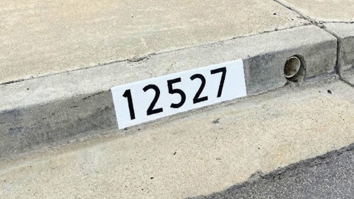 Black number 12527 painted on a white curb marker next to a drainage pipe opening in the curb