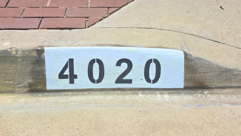 Black number 4020 painted on a white background curb address marker beside a brick paver walkway